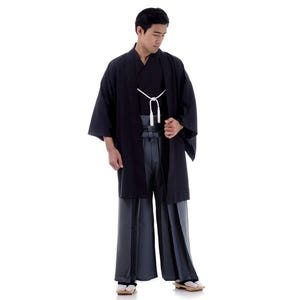 May include: A man wearing a black kimono with a white sash and grey trousers. He is standing with his hands clasped in front of him.