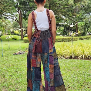 May include: A woman wearing a white tank top and a pair of patchwork wide-leg overalls. The overalls are made of different fabrics and colors, including blue, green, red, and brown. The woman is standing in a grassy area with trees in the background.