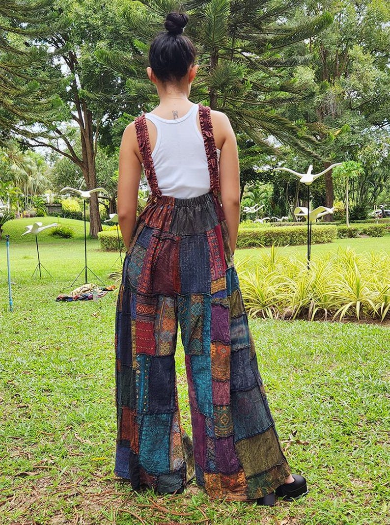 May include: A woman wearing a white tank top and a pair of patchwork wide-leg overalls. The overalls are made of different fabrics and colors, including blue, green, red, and brown. The woman is standing in a grassy area with trees in the background.