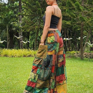 May include: A woman wearing a black tank top and patchwork pants with a variety of colors and patterns. The pants are wide-legged and flowy. The woman is standing in a grassy area with trees in the background.