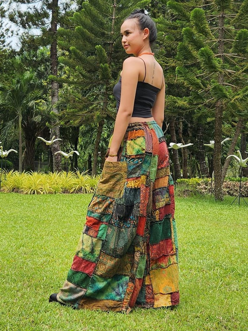 May include: A woman wearing a black tank top and patchwork pants with a variety of colors and patterns. The pants are wide-legged and flowy. The woman is standing in a grassy area with trees in the background.
