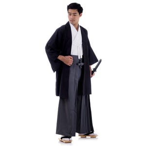 Op de afbeelding: Een man die een zwarte kimono draagt met een wit onderkleed en een zwaard. Hij staat met zijn hoofd gebogen en houdt het zwaard in zijn rechterhand.