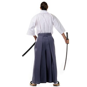 Op de afbeelding: Een persoon draagt een witte kimono en een grijze hakama broek, met een katana zwaard in de rechterhand.