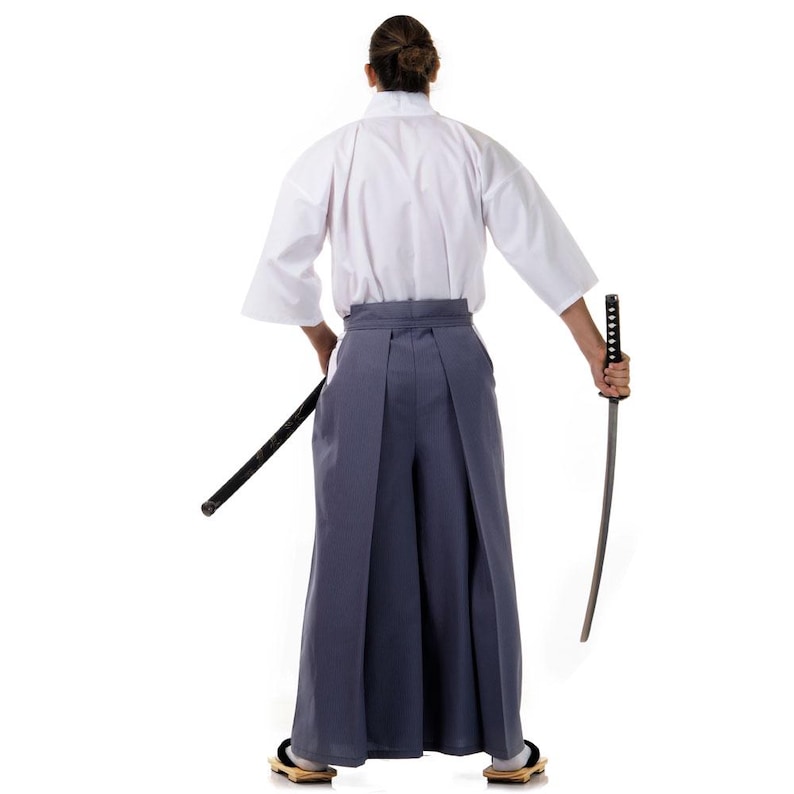 Op de afbeelding: Een persoon draagt een witte kimono en een grijze hakama broek, met een katana zwaard in de rechterhand.