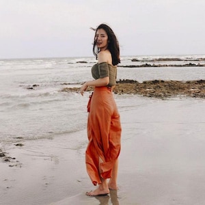 Op de afbeelding: Een vrouw in een groen, off-shoulder top en een oranje wijde broek staat op een zandstrand.