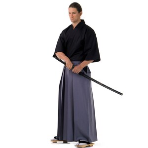 May include: A man wearing a black kimono and grey hakama trousers holds a black katana sword. He is standing against a white background.