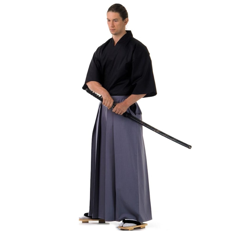 May include: A man wearing a black kimono and grey hakama trousers holds a black katana sword. He is standing against a white background.