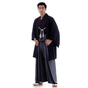 May include: A man wearing a black kimono with a white sash and a grey hakama. He is standing with his hands on his hips.