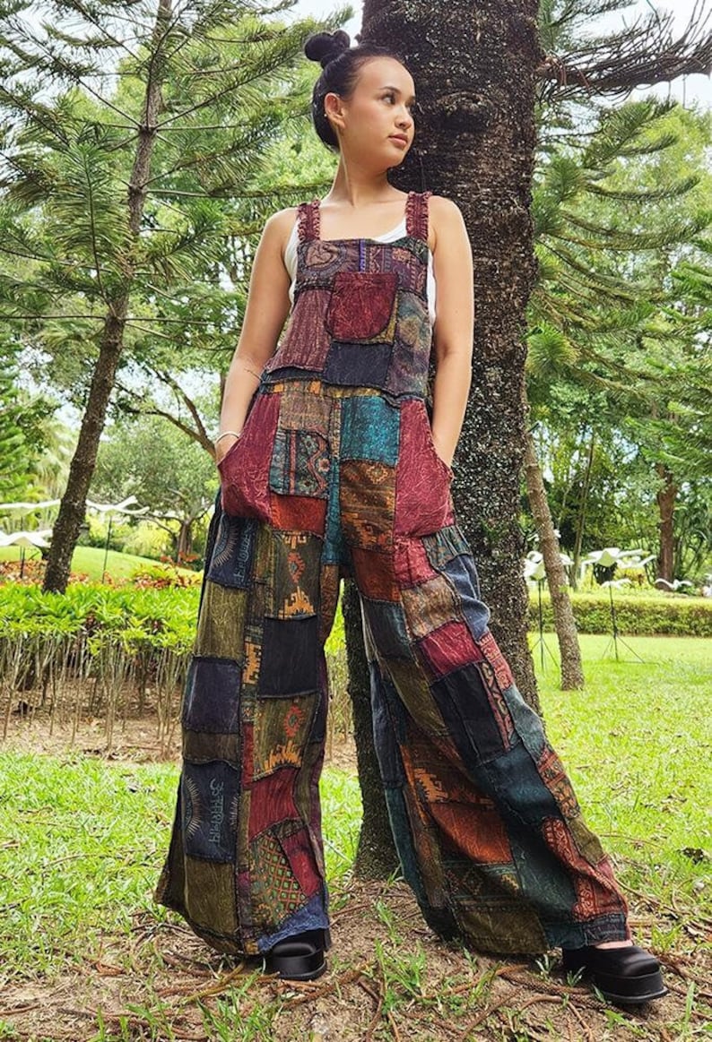 May include: A woman wearing a patchwork jumpsuit with a variety of colors and patterns. The jumpsuit has wide legs and a relaxed fit. The woman is standing in a grassy area with trees in the background.