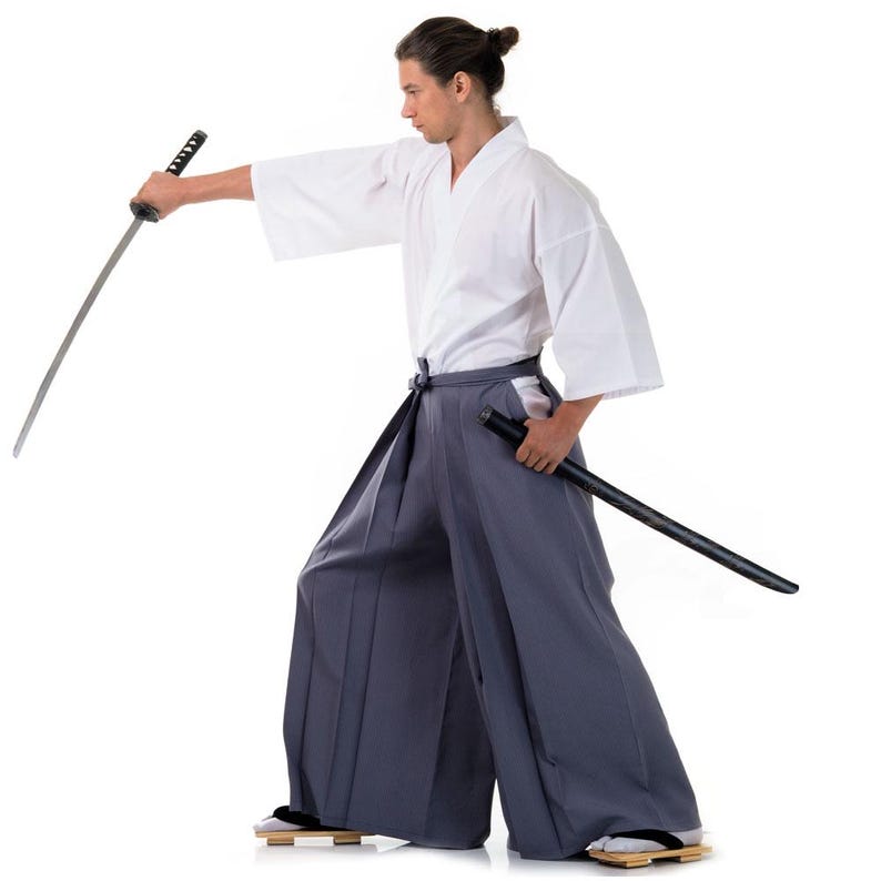 Op de afbeelding: Een persoon in een wit martial arts uniform en grijze broek houdt een zwart katana-zwaard in zijn rechterhand. Het zwaard wordt vastgehouden in een traditionele greep, met de kling naar voren gericht. De persoon staat op een witte achtergrond.