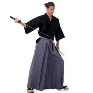 May include: A person wearing a black kimono and grey hakama trousers holds a silver sword in their right hand. They are standing against a white background.