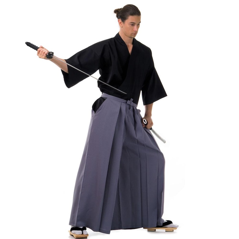 May include: A person wearing a black kimono and grey hakama trousers holds a silver sword in their right hand. They are standing against a white background.