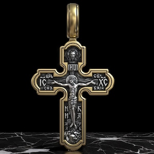May include: An Orthodox cross pendant in gold and silver tones. The cross depicts Jesus Christ in detail, with inscriptions in an old Slavic script. The pendant has a loop for a chain and is set against a dark background.