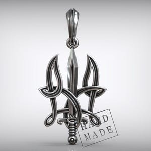 May include: Silver pendant featuring a stylized trident design with a sword at its center. The pendant has a loop at the top for a chain and a small tag that reads "HAND MADE". The pendant is made of a shiny, metallic material.