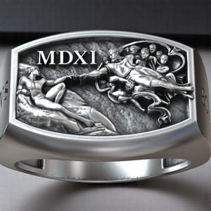 Può includere: Anello d'argento con un rilievo dettagliato della Creazione di Adamo, e il testo "MDXI". L'anello ha una fascia larga e una finitura lucida, che mette in mostra l'intricata opera d'arte. Gioielleria.