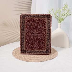 Peut inclure: Un étui rectangulaire avec un motif de tapis traditionnel dans des tons bordeaux, noir et or. L'étui est posé sur un dessous de plat rond et tissé. Un vase blanc avec des fleurs est à l'arrière-plan.