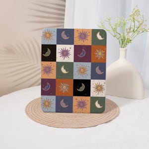 May include: A square, decorative item featuring a repeating pattern of suns and crescent moons in various colors. The design is arranged in a grid of squares, with each square containing a different combination of celestial elements. The color palette includes shades of orange, purple, and blue.