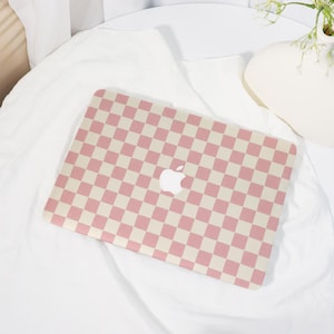 May include: A laptop with a pink and cream checkerboard pattern. The laptop has a white Apple logo in the center. The laptop is resting on a white surface.