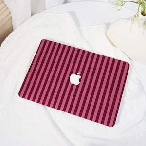 May include: A burgundy and pink striped laptop skin with the Apple logo in white. The skin is rectangular and covers the top of the laptop. The background is a white sheet.