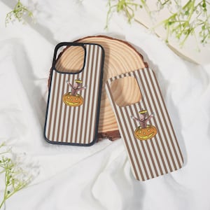 May include: Two phone cases with a brown and white striped pattern. One case has a black border. Each case features a cartoon monkey emerging from a carved pumpkin. The cases are on a wooden slice.