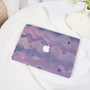 May include: A laptop case with a vibrant abstract design. The case displays a wavy pattern in shades of purple, pink, and blue, with the Apple logo. The case is designed to fit a laptop.