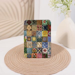 May include: A square decorative object with a mosaic tile design. The tiles feature various floral, geometric, and sunburst patterns in a range of colours including blue, yellow, red, and green. The object is placed on a round, woven mat.