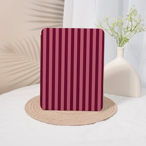 May include: A tablet case with a vertical striped pattern in shades of burgundy and pink. The case is resting on a round, woven placemat. A white vase with flowers is in the background.