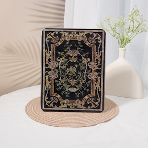 Peut inclure: Un panneau décoratif rectangulaire noir avec un motif floral et d'oiseaux orné. Le panneau présente une bordure dorée et est posé sur un tapis tissé beige. Un vase blanc avec des fleurs est à l'arrière-plan.