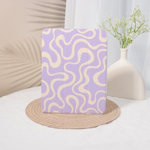Könnte beinhalten: Eine lavendelfarbene Tablethülle mit einem cremefarbenen Wellenmuster. Die rechteckige Hülle liegt auf einem runden, gewebten Platzset. Eine weiße Vase mit Blumen befindet sich im Hintergrund.