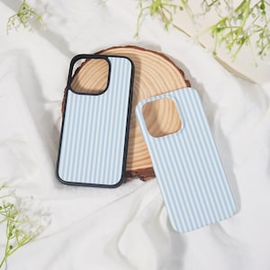 May include: Two phone cases with a light blue and white striped pattern. One case has a black border. The cases are resting on a wooden slice and white fabric, with greenery in the background.