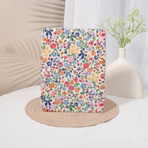 May include: A rectangular case with a colorful floral pattern featuring red, pink, blue, and yellow flowers on a cream background. The case is resting on a round, woven placemat. A white vase with flowers is in the background.