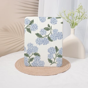 May include: An off-white tablet case featuring a repeating pattern of blue hydrangea flowers and green leaves. The case is displayed on a round, woven placemat. A white vase with flowers is in the background.