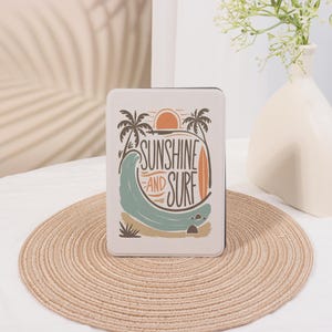 May include: A white book cover with a graphic design of palm trees, a sunset, a wave, and a surfboard. The text on the cover reads "Sunshine and Surf".
