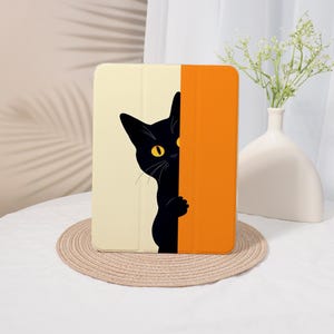 May include: A tablet case featuring a black cat peeking from behind a cream and orange panel. The cat has yellow eyes and a playful pose. The case is on a woven mat.