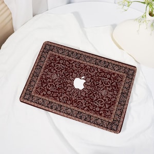 May include: A laptop with a decorative cover featuring a Persian rug design. The cover is predominantly burgundy with intricate floral patterns in beige and black. The Apple logo is visible in white.