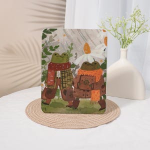 Könnte beinhalten: Eine rechteckige Tablethülle mit einer Aquarellillustration von zwei Fröschen, die im Regen spazieren gehen. Ein Frosch trägt einen roten Schal und eine karierte Jacke, der andere einen orangefarbenen Schal und einen braunen Mantel, umgeben von grünem Laub und weißen Blumen.