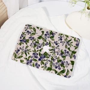 May include: A laptop case featuring a watercolor design of blueberries and green leaves on a light beige background. The case has a cutout for the Apple logo and is placed on a white surface.
