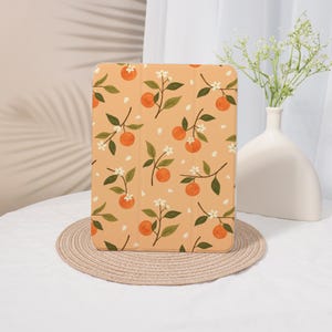 May include: Tablet case featuring a repeating pattern of orange citrus fruit, green leaves, and white blossoms on a peach background. The case is rectangular and rests on a round, woven placemat.
