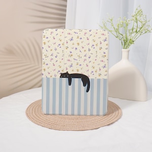 May include: A rectangular tablet case featuring a split design. The top section has a cream background with a small purple and yellow floral pattern. The bottom section has light blue and white vertical stripes with a black cat illustration.