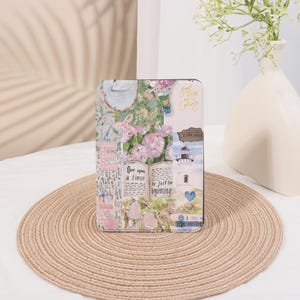 May include: A decorative case featuring a collage of floral patterns, a lighthouse, and the text "One upon a time is just the beginning." The case has a pastel color scheme with pink, green, and white accents. The case is rectangular and appears to be for a tablet or e-reader.