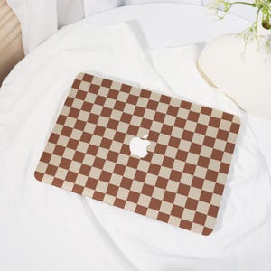 May include: A laptop featuring a brown and beige chequered design. The Apple logo is visible in the centre. The laptop is placed on a white surface.