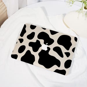 May include: A laptop with a cow print design. The laptop has a beige background with large black spots. The Apple logo is visible in the center. The laptop is placed on a white surface.