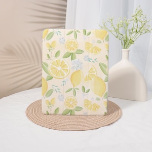 May include: A light-colored tablet case with a lemon pattern. The case features illustrations of lemons, lemon slices, and leaves in shades of yellow and green. The case is on a round, woven placemat.