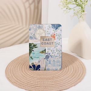 May include: A rectangular photo frame with a collage of coastal images, including seashells, a lobster, and beach scenes. The text "EAST COAST" is centered in the frame. The frame is on a round, woven placemat.