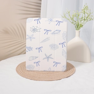 May include: A white tablet case featuring a pattern of blue seashells, starfish, and bows. The case is resting on a round, woven placemat. A white vase with greenery is in the background.