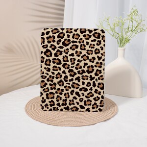 May include: A rectangular tablet case featuring a leopard print design in shades of brown and black. The case is placed on a round, woven placemat. A white vase with flowers is in the background.