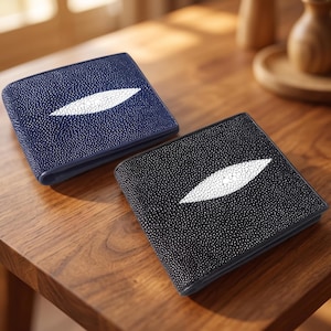 Op de afbeelding: Twee bifold portemonnees met een textuur van roggenhuid. De ene portemonnee is donker marineblauw en de andere is zwart. Elke portemonnee heeft een opvallend, langwerpig wit ruitvormig patroon in het midden van de voorzijde.
