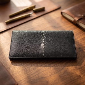 May include: A rectangular black wallet featuring a textured stingray skin surface with a distinct, lighter-coloured central spine pattern composed of small, rounded beads running vertically down the middle of the exterior panel.