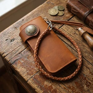 Può includere: Un portafoglio in pelle color cuoio con un bottone concho circolare in ottone raffigurante il profilo in rilievo di una figura nativa americana. Il portafoglio include un cordino in pelle intrecciata con una clip girevole in metallo.
