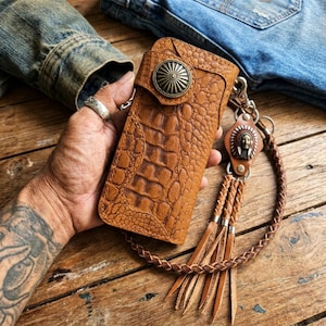 May include: A tan leather wallet with a textured crocodile pattern, featuring a circular metal concho snap closure. Attached is a braided leather lanyard with a metal charm depicting a person in a headdress and three leather fringe tassels.